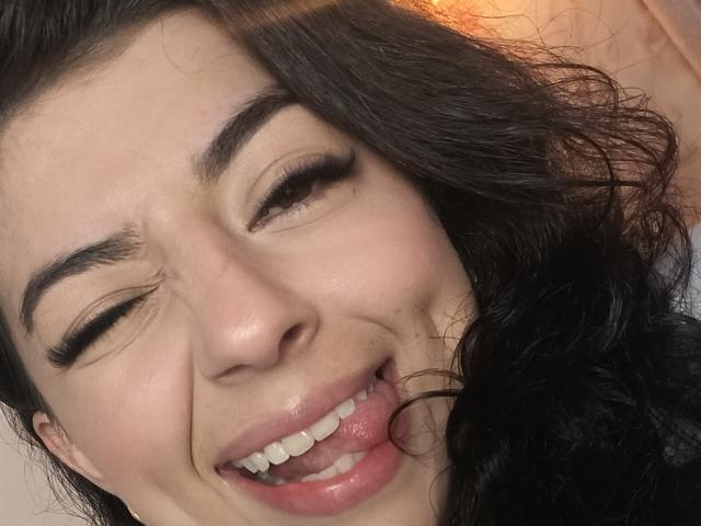 AnntonellaWatson's face with wavy black hair, closed eyes, and a playful smile showing a small bit of her pink tongue and white teeth. Her eyebrows are raised slightly.