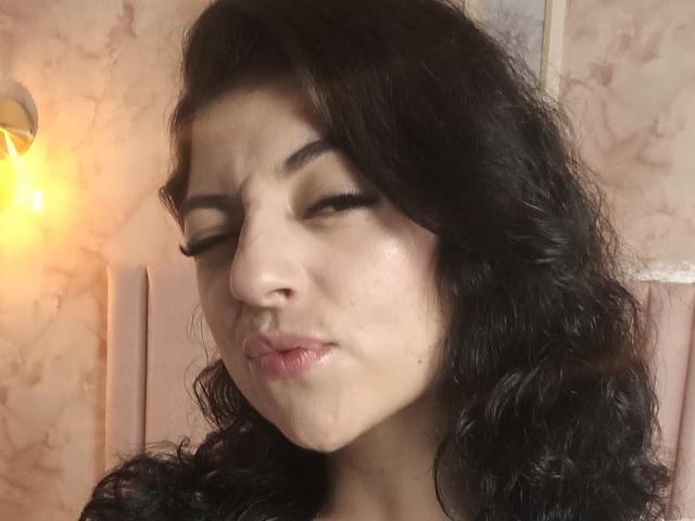 AnntonellaWatson has dark curly hair, closed eyes, and puckered lips in a pouty expression. She's a pale-skinned woman with visible eyelashes and a slight sheen on her face.