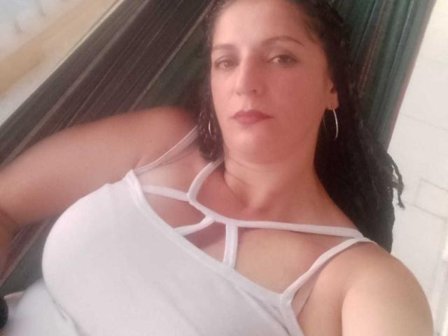 MadameScarlett es una mujer de piel media con cabello largo y castaño oscuro. Lleva un top blanco con tirantes finos que realza sus grandes senos. Su expresión facial es neutra, y lleva aros grandes en ambas orejas. La fotografía está ligeramente borrosa, enfocándose en la parte superior de su cuerpo. Tiene un ligero rubor en las mejillas, y la fuente de luz es suave, creando sombras sutiles. Está mirando ligeramente hacia la izquierda..