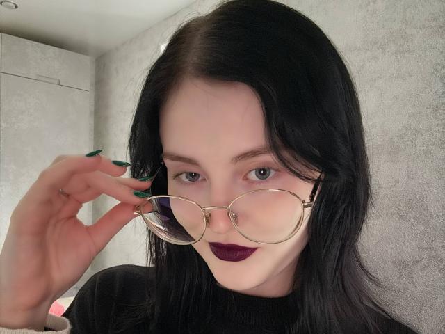 MakedaRiedell with dark hair, purple lipstick, green nails, and gold glasses, holding them with her right hand, showing off her cute face.