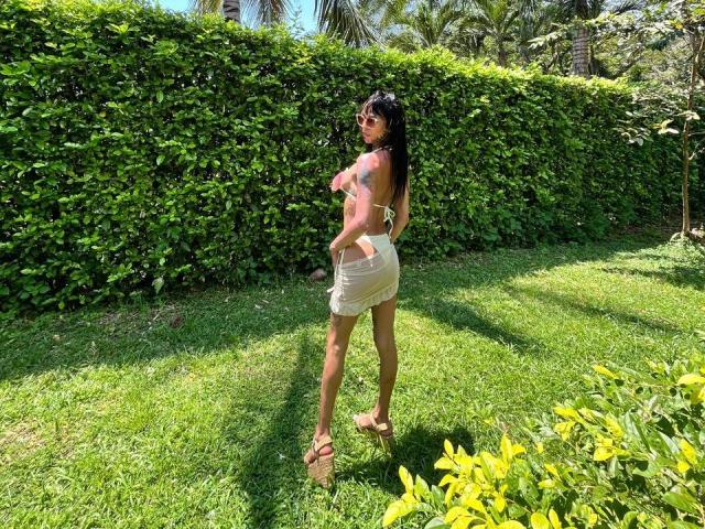 AnneKisss stands in a sunny garden, facing away from the camera. She has long black hair, tan skin, and is wearing a white, semi-transparent dress that reveals her back and a small part of her buttocks. She's also wearing pink, strappy sandals. The green grass and yellow plants in the foreground contrast with the dense, green hedge in the background. Palm trees are visible in the distance. The sunlight casts shadows on the grass.