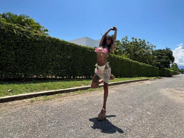 AnneKisss stands on a sunny road, arms raised, wearing a pink floral bikini top and a sheer floral skirt. She has long dark hair, a tattoo on her right thigh, and is wearing tan wedge sandals. The bright blue sky and lush green hedge are in the background. The road is gray with a slight shadow under her. Her right leg is bent, and she's looking to the side.