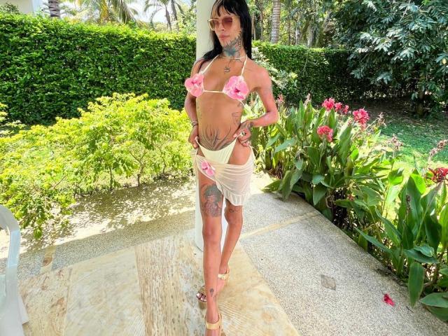 AnneKisss stands in a sunny garden, wearing a pink floral bikini top and a sheer, white wrap skirt. She has multiple tattoos on her arms and legs, dark hair, and sunglasses. Her tan skin contrasts with the greenery and pink flowers around her. She's in high-heeled sandals, with her hands on her hips. Bright natural light emphasizes her confident stance and the floral pattern on her bikini.