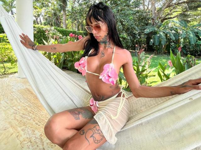 AnneKisss sits in a white hammock, wearing a pink flower bikini top and a sheer beige skirt. She has long black hair, pink-tinted sunglasses, and multiple tattoos on her arms and thighs. Her skin is tan, and she has a slim, toned physique. She's holding the hammock with her left hand, her right arm resting on her knee. The hammock is suspended in a lush, green garden with trees and plants in the background. The texture of the hammock is smooth, and the garden is bright with sunlight.