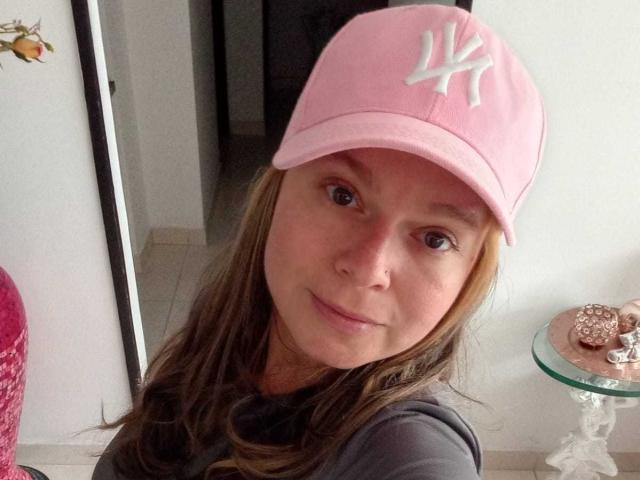 PamelaColin with long brown hair, wearing a pink NY Yankees cap, smiling at the camera, grey shirt, nose ring, small round brown eyes, and a glass table with cookies.