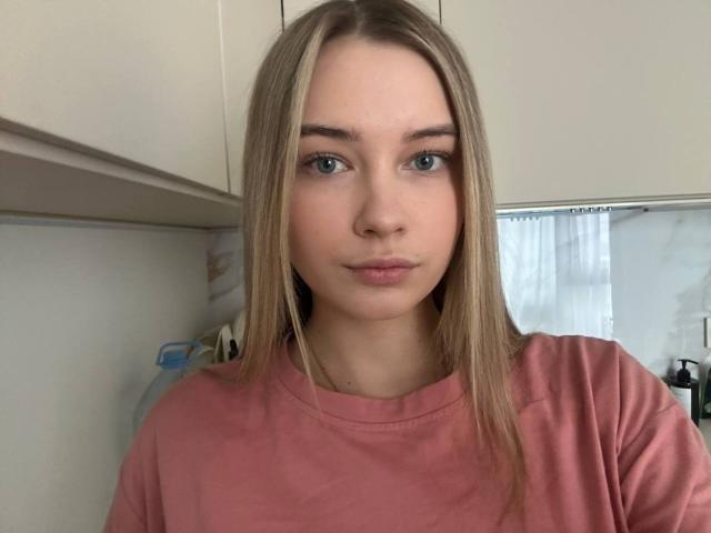Portrait of LavondaVanors, a young woman with straight, blonde hair, blue eyes, and fair skin. She wears a pink, short-sleeved shirt. Her expression is neutral, with slightly parted lips. The background is a simple, indoor setting with white cabinets and a partially visible curtain. The image is sharp with even lighting. Her eyebrows are thin and well-groomed, and she has a natural, minimal makeup look. The overall composition is centered, with LavondaVanors' face taking up most of the frame.