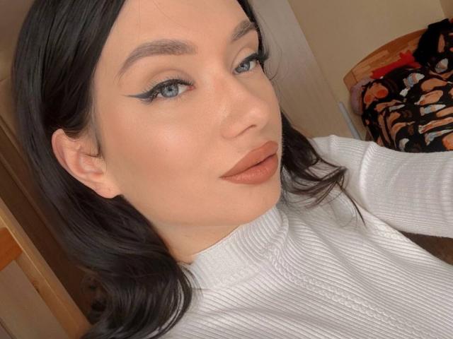 AlessandraShoe has blue eyes, black eyeliner wings, and brown lipstick. She's wearing a white ribbed turtleneck, with dark wavy hair, and a neutral, smooth face.