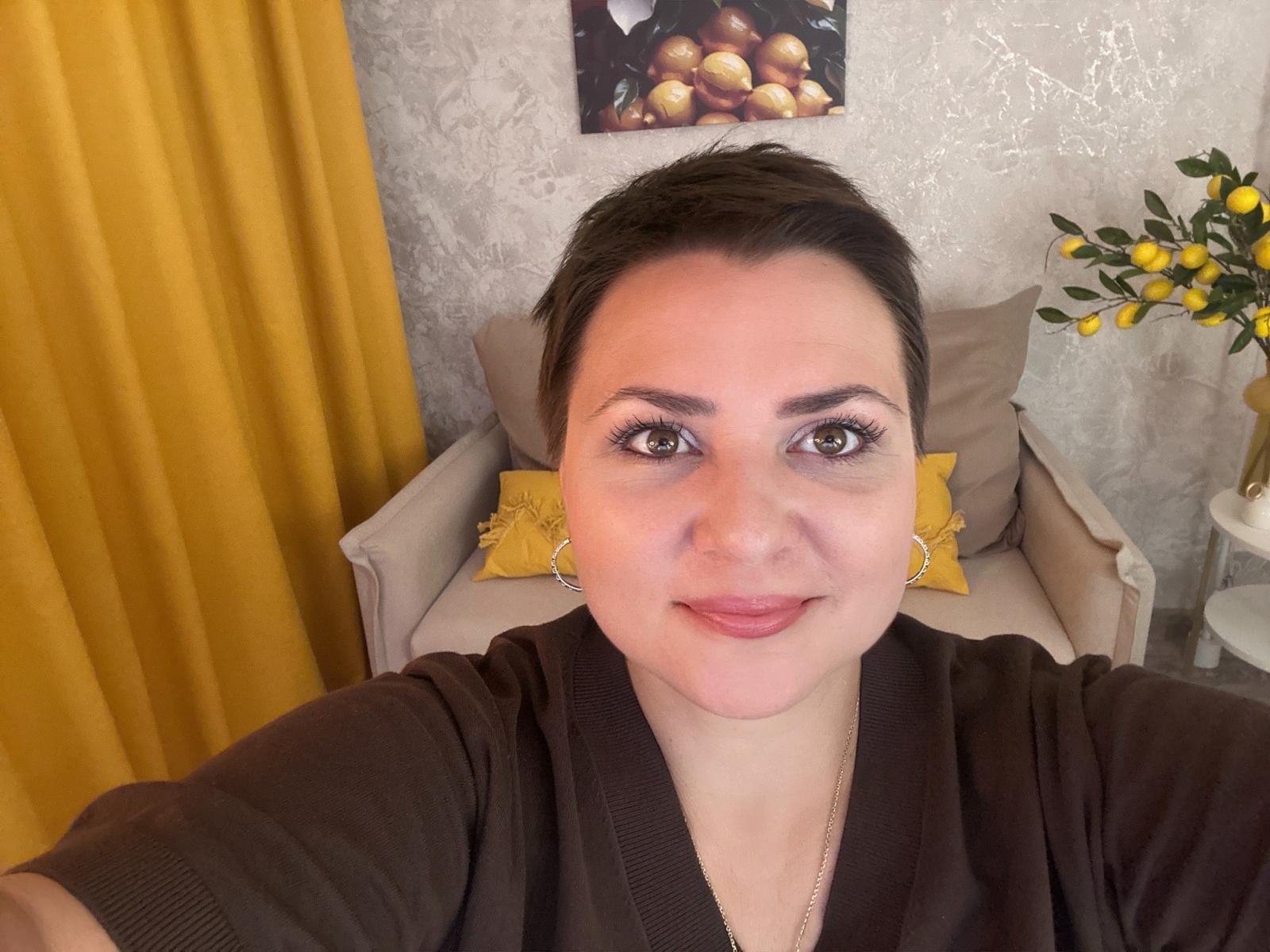 AnnaCorlane has a short, dark brown pixie haircut, light skin, and brown eyes with subtle eyeliner. She wears a dark brown zip-up hoodie and gold hoop earrings. She's smiling slightly, looking directly at the camera. The background includes a yellow curtain, a beige armchair with a yellow pillow, and a decorative lemon painting on the wall. A small white table with yellow lemons is on the right. The texture of the wall is slightly rough. The image has a warm, natural light.