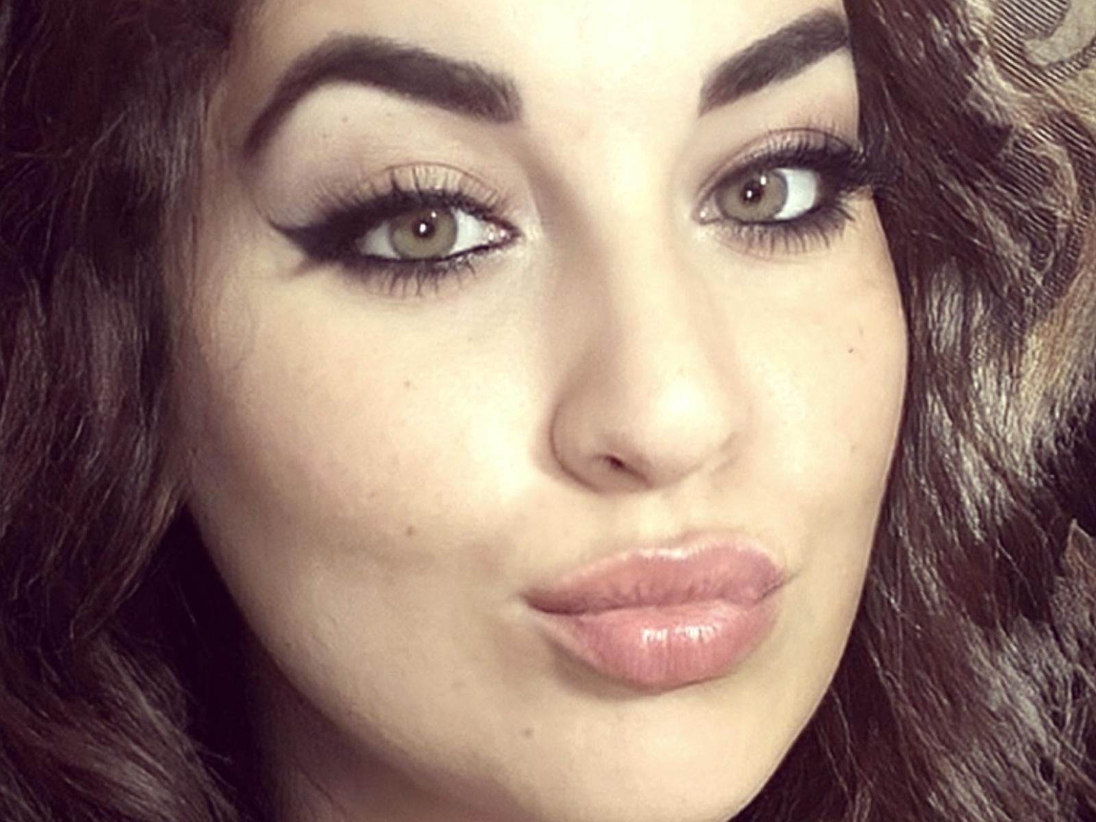 Yzabella69's face, featuring green eyes, thick black eyebrows, long eyelashes, and glossy pink lips. She has wavy dark brown hair and a light skin tone.
