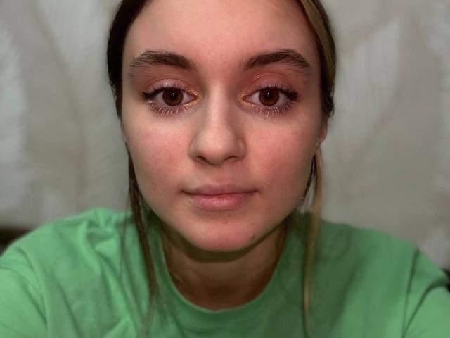 Close-up of TerisaCountess. She has fair skin, brown eyes with light brown eyeliner, and slightly arched eyebrows. Her brown hair is pulled back with a few strands falling forward. She wears a green shirt. The lighting is even, highlighting her face. The background is blurred and indistinct. Her expression is neutral, with slightly parted lips. The image is sharp with a realistic texture.