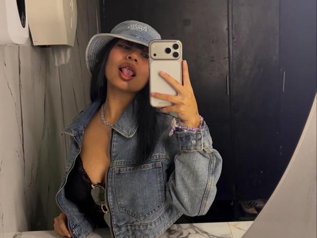 VannesaSaenz takes a mirror selfie in a bathroom. She has long black hair, wears a blue denim hat with white text, and a matching denim jacket over a black lace bra. Her tongue is out, and she holds a white smartphone with a red case. She has a silver chain necklace and a pink scrunchie on her right wrist. The image has a casual, street-style vibe with a focus on her confident, playful expression. The texture of the denim and the lace bra contrasts with her smooth skin.