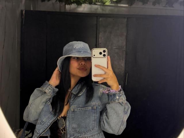 VannesaSaenz takes a mirror selfie. She's wearing a blue denim jacket, a matching bucket hat with "Los Angeles" text, and has long black hair. She holds a white iPhone with a three-camera setup in her right hand, and her left hand touches the hat. She has a slight smile and is looking at the phone. The background is dark with a black cabinet and some greenery above. The image has a casual, urban style.