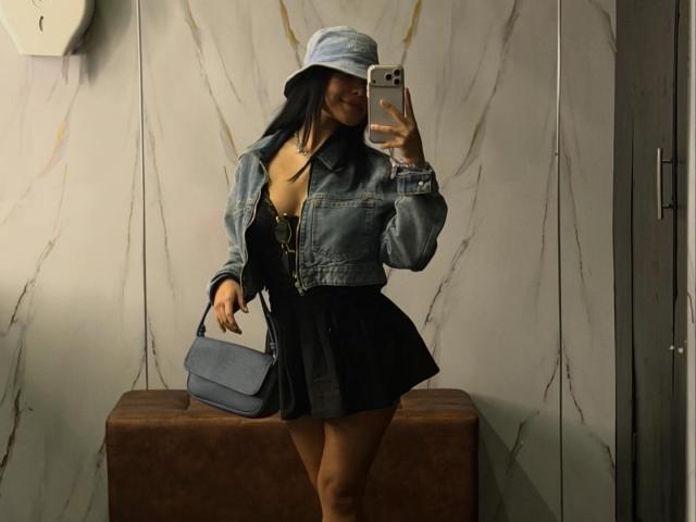 VannesaSaenz takes a mirror selfie in a modern, marble-walled room. She wears a blue denim jacket over a black, A-line dress, with a light blue bucket hat covering her face. She holds a gray, leather handbag in her right hand. Her long, dark hair is partially visible. The brown, cushioned bench she stands in front of is positioned against the marble wall. The image has a casual, contemporary style.