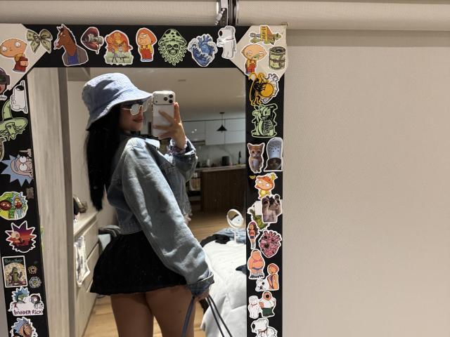 VannesaSaenz takes a mirror selfie in a room. She's wearing a blue bucket hat, a light denim jacket, and a black mini skirt. Her long black hair is partly visible. The mirror is decorated with colorful stickers, including cartoon and pop culture characters. She's holding an iPhone with a pink case. Her reflection shows her back and a partial side view. The room has wooden flooring and a bed with white sheets.