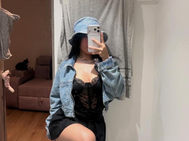 VannesaSaenz takes a mirror selfie. She wears a denim bucket hat, unbuttoned denim jacket, and black lace corset with a black skirt. Her phone, a silver iPhone, partially covers her face. She has long black hair and a silver choker. The image has a casual, modern style with a focus on streetwear. The background includes a beige sofa and a black cat. The wooden floor adds warmth to the scene.