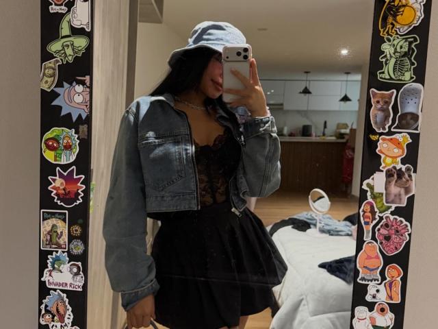 VannesaSaenz stands in front of a mirror, taking a selfie. She wears a light blue bucket hat, a black lace dress with a deep neckline, and a denim jacket. Her long black hair is visible under the hat. The mirror is decorated with colorful stickers on both sides. She holds a white smartphone with her right hand. The room has a bed with a white blanket and a wooden floor. The image is a realistic photograph.