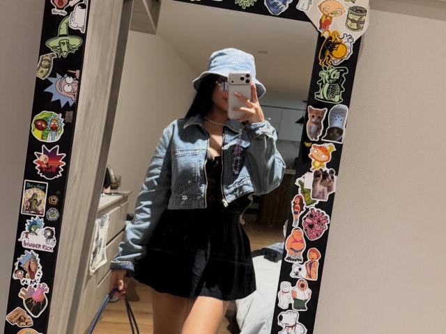 VannesaSaenz stands in front of a mirror, taking a selfie. She wears a light blue denim jacket with the sleeves rolled up, over a black mini dress. She also has on a light blue bucket hat and holds a white smartphone in her right hand. The mirror's black frame is decorated with colorful stickers, including characters and objects. Her long black hair is partially visible. The room has a wooden floor and white walls.