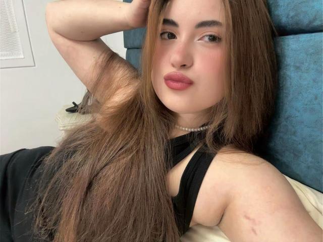 Close-up of VicluX, a young woman with long, straight brown hair, lying on a blue, patterned pillow. She has fair skin, subtle makeup, and pinkish lips. She wears a black sleeveless top and a pearl necklace. Her right arm is raised, with her hand resting behind her head. The image has soft lighting, highlighting her relaxed expression. A small scar is visible on her right arm. The composition focuses on her face and upper body.