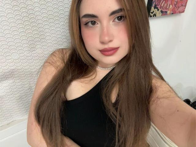 VicluX has long, straight brown hair, wearing a black one-shoulder top. She has fair skin, pink blush on her cheeks, and dark, well-defined eyebrows. Her lips are painted a muted red. She wears a small pearl choker. The background has a white, textured pattern on the left and colorful art on the right. The image is a selfie, focusing on her upper body. Her expression is neutral with a slight smile.