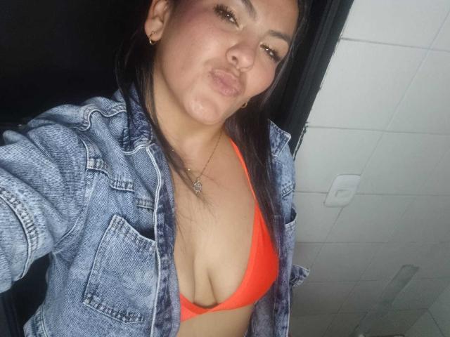 ScarlettOxford, a medium-brown-skinned woman with straight black hair, wears a blue denim jacket over a bright orange bikini top. She has a pouty expression, small gold earrings, and a silver necklace. Her cleavage is prominent. The image is a close-up selfie, emphasizing her upper body. The background is indistinct.