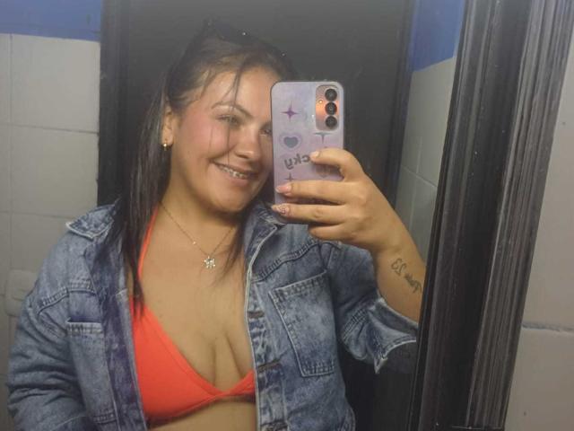 ScarlettOxford with wet brown hair, wearing a red bikini top showing her tits, denim jacket, smiling at mirror selfie, holding purple phone with purple stickers, tattoo on right wrist.
