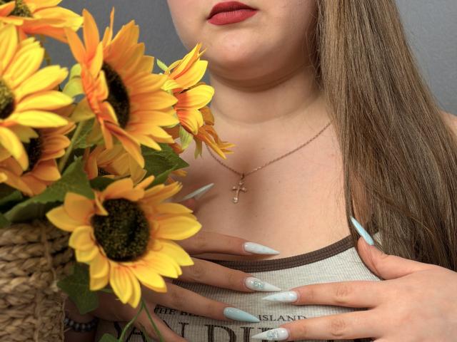 Close-up of AvaNiss holding sunflowers. AvaNiss has long, straight brown hair and wears red lipstick. She's wearing a white tank top with brown trim and a gold cross necklace. Her hands have long, light blue nails with white patterns. Sunflowers in the foreground are vibrant yellow with dark green centers and bright orange petals. AvaNiss's right hand is gently holding the sunflowers while her left hand rests on her chest. The image has a soft, natural light.