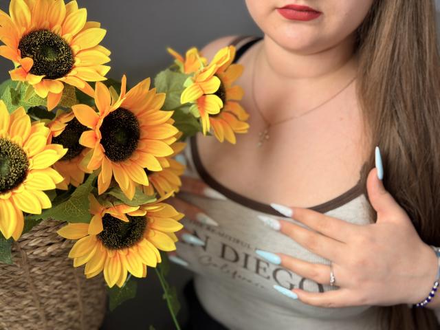 Close-up of AvaNiss with sunflowers. She has light skin, red lips, and long brown hair. She wears a beige tank top with brown trim and a necklace. Her right hand with long, white, and blue manicured nails touches her chest. Sunflowers with yellow petals and dark centers are in a woven basket on the left. Her left hand rests on her chest. The image has a natural, realistic style. Her ring finger has a small ring.