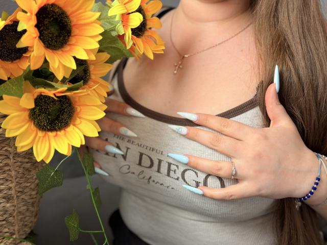 Close-up of AvaNiss with sunflowers in the foreground. She has long, straight brown hair, wearing a beige ribbed tank top with brown trim and "The Real San Diego" text. Her hands, with long, light blue nails, cover her chest. She wears a gold necklace, a ring, and a blue bead bracelet. The sunflowers are vibrant yellow with dark brown centers, and green leaves. The texture of the tank top contrasts with the sunflower petals. AvaNiss's skin is light. The image is sharp with natural lighting.