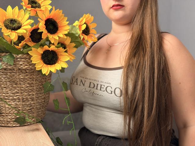 AvaNiss with long brown hair, red lipstick, and a tank top showing her big boobs, holding a basket of sunflowers, her hand on her dark jeans.