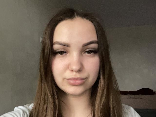 JackiShular has long brown hair, light skin, and full lips with a subtle smile. She has brown eyes, dark eyebrows, and is wearing a white shirt.