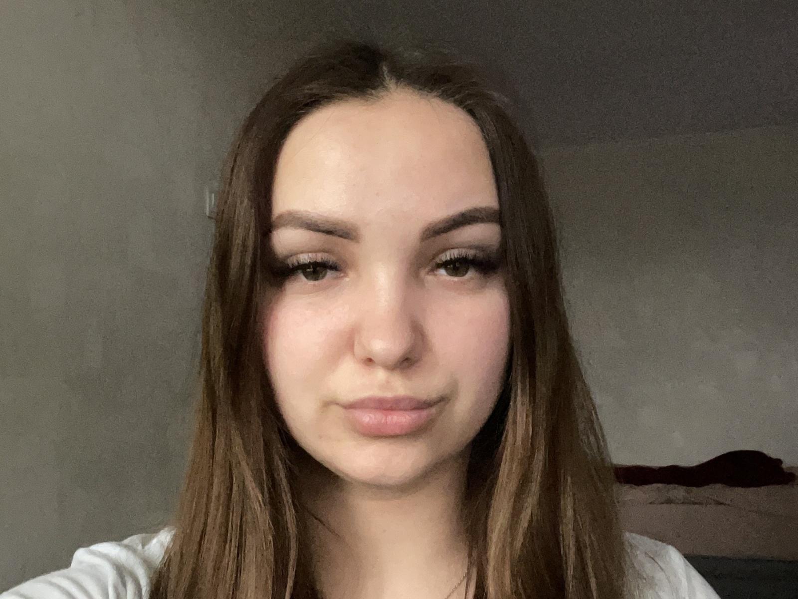 JackiShular has long brown hair, light skin, and full lips with a subtle smile. She has brown eyes, dark eyebrows, and is wearing a white shirt.