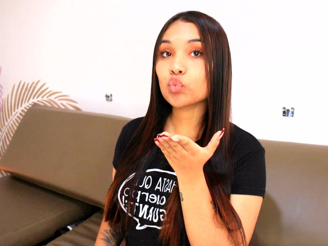 BelleLane with long brown hair, pouting lips, and black shirt, holding hand up, showing red nails, and looking at camera. Brown sofa and white wall in background.