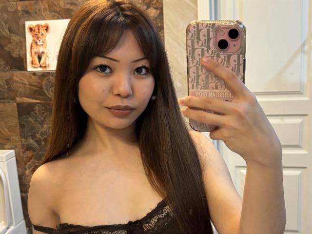 Joetta Bealer, con cabello castaño largo, tomando una selfie con su teléfono, vistiendo una blusa negra de encaje, mostrando sus senos, y con una sonrisa sutil..