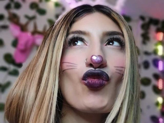 Close-up of Kaiix with blonde hair, wearing purple glittery lipstick and a pink heart-shaped nose. Kaiix has pink whisker-like marks on her cheeks. She looks upward with her eyes slightly squinted. Kaiix's eyelashes are long and dark. The image has a blurred, colorful background with green leaf patterns and pink objects. Kaiix's makeup is bold and playful, contrasting with her natural hair. The texture of her lipstick is shiny and reflective. Her nose piercing is small and silver. The composition focuses on Kaiix's facial expression and makeup details.