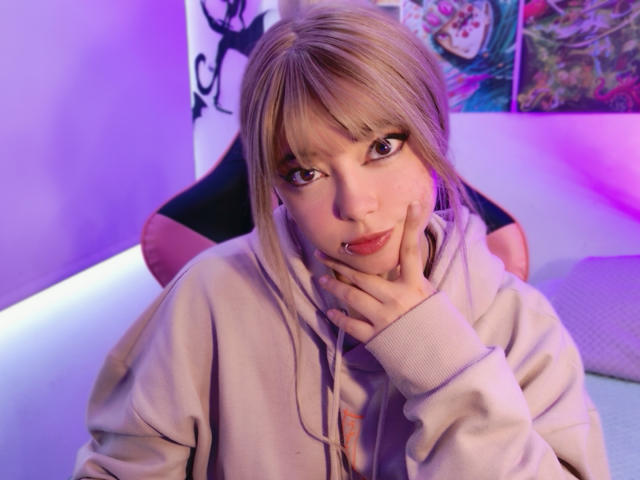 Arieyaara, a young woman with fair skin and light blonde hair, stares directly at the camera. She has bangs, large dark eyes with long eyelashes, and a small nose ring on her right nostril. She wears a beige hoodie with her left hand touching her cheek. The purple and blue lighting in the background contrasts with the soft white and pink elements. Her expression is calm and slightly curious. The image focuses on her upper body and face, with a slight overexposure on her face.