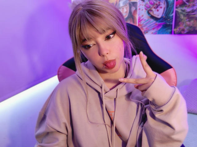 Arieyaara has light blonde hair with bangs, wearing a gray hoodie, and is making a pouty face with her tongue out. She's holding up a finger to her lips. Her eyes are half-closed. The background is purple-lit with colorful anime posters. The image has a soft, slightly blurred effect with warm, pinkish lighting. The chair she's sitting on is black with pink accents. The composition focuses on Arieyaara's face and hand.