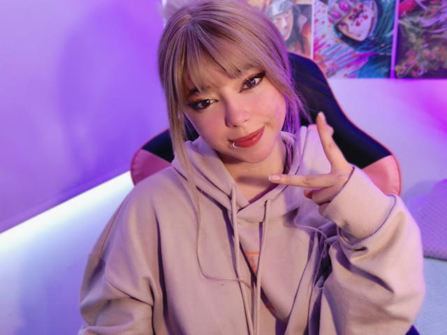 Arieyaara, a young woman with light blonde hair and bangs, sits in a pink and black chair. She wears a light gray hoodie and has a pink lip ring. Her right hand is raised, making a "shoo" gesture with her fingers. Her makeup includes dark eyeliner and red lipstick. The background is blurred with purple and pink lights, and anime posters are visible. The image has a soft, digital filter effect.