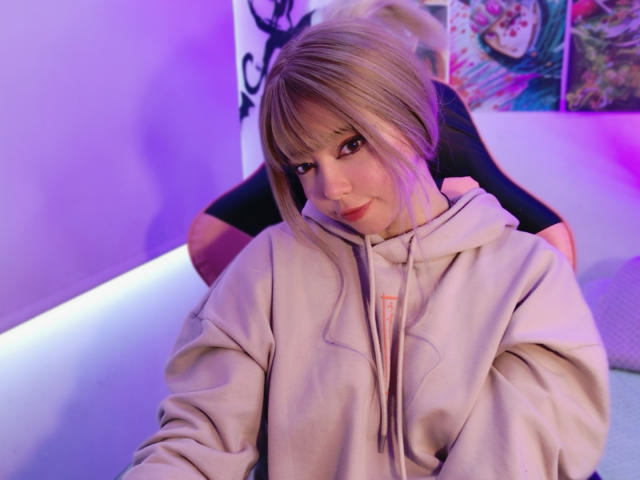 Arieyaara, a blonde woman with straight bangs, sits in a relaxed position, looking at the camera with a slight smile. She wears a beige hoodie with a pink logo on the chest. Her makeup is subtle, with pink lipstick. The purple and blue lighting casts a soft glow on her face. In the background, a black and white illustration is partially visible. The composition is casual and slightly angled. The texture of the hoodie is soft and slightly loose. Her expression is relaxed and slightly playful.