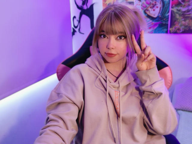 Arieyaara, a young woman with light brown hair in bangs, sits in a chair. She wears a beige hoodie with a pink zipper. She makes a peace sign with her right hand. Her brown eyes look at the camera. The room has purple and blue lighting, with a colorful poster on the right and a black silhouette on the wall. The image is clear, with a soft focus on Arieyaara. She has a cute, slightly smiling expression. Her hair is straight and parted in the middle.