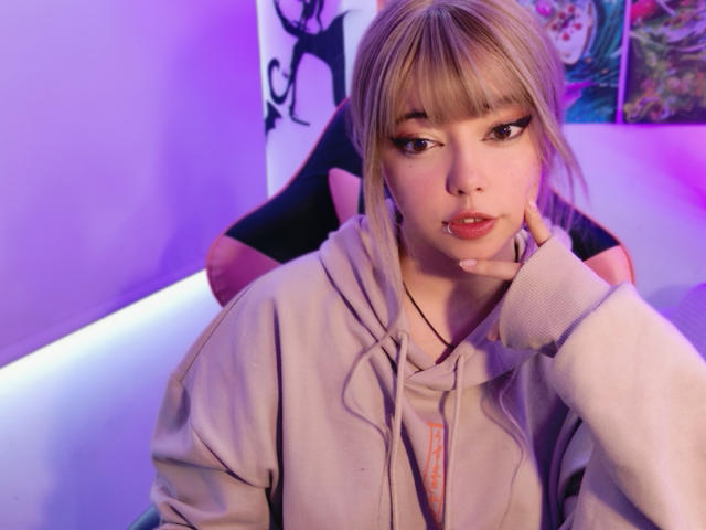 Arieyaara, a young woman with light skin and blonde hair, sits in a black and pink chair. She has full lips, dark eyeliner, and is wearing a grey hoodie. Her right hand rests on her cheek. The image is lit with purple and pink lighting. Her expression is neutral, and she has a small nose ring. The background is blurred, featuring abstract art. The photo has a soft focus on Arieyaara's face.