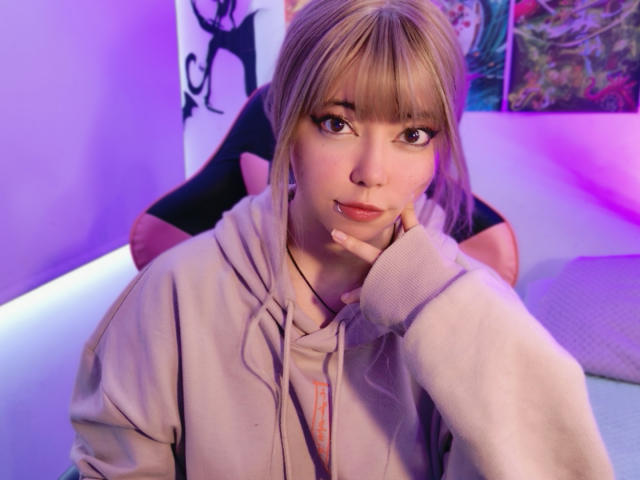 A photograph of Arieyaara, a young woman with blonde hair and bangs, wearing a beige hoodie. She's sitting in a black chair, her left hand resting on her cheek. Her brown eyes look directly at the camera. Her lips are slightly parted. The background has purple and pink lighting with abstract art on the walls. The image has a soft, warm glow with visible texture on her hoodie and the chair. Her facial expression is calm and slightly curious.