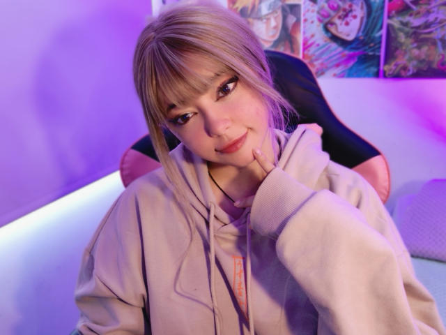 Arieyaara, a young woman with fair skin and blonde hair in a side braid, sits in a black chair with pink accents. She wears a light beige hoodie with orange drawstrings. Her right hand touches her chin, and she smiles slightly. The background features colorful anime posters. The lighting is purple, creating a soft glow on her face. The image is a photograph with a casual, relaxed composition.