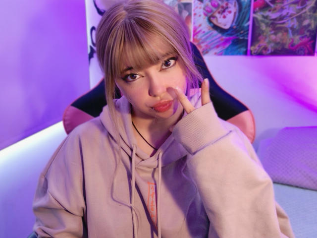 Arieyaara with blonde hair and bangs sits in a chair, pouting with her index finger on her cheek. She wears a beige hoodie, and the background has purple and blue lighting with colorful posters. Her brown eyes are slightly squinted, and she has light skin. The chair is black with pink accents. The image has a soft, colorful aesthetic with a mix of purple and blue lighting. The focus is on Arieyaara's expressive face and casual posture.