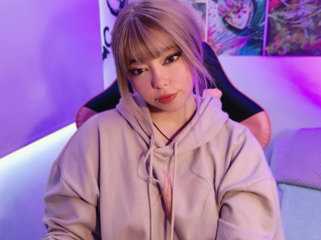 Arieyaara, a young woman with light blonde hair in a straight bob with bangs, sits in the foreground. She wears a light gray hoodie with a small orange design near the chest. Her dark eyebrows and eyeliner contrast with her pinkish-red lipstick. She has a subtle nose ring on her right nostril. The background is blurred with purple and pink lighting. Her light skin is visible, and her expression is neutral. The composition focuses on her upper body and face.