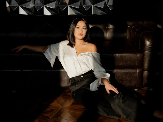 AlyceSkagen is seated on a dark brown leather couch, illuminated by a strong light source. She has medium-length, straight black hair and a light olive skin tone. She wears a white, off-the-shoulder blouse with loose sleeves and black high-waisted pants. Her right arm is extended along the couch, while her left hand rests on her lap. The geometric patterned wall in the background is partially visible. The wooden floor has a herringbone pattern. Her red nails stand out against the dark background. The light creates sharp shadows, emphasizing her relaxed posture and confident expression.