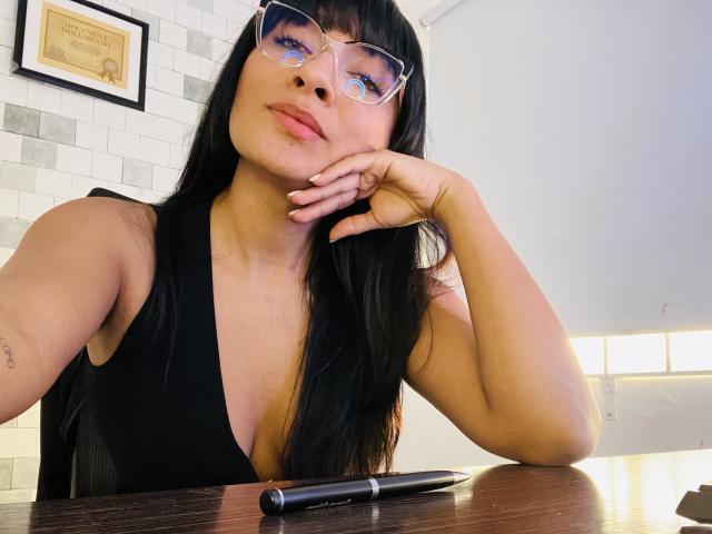 AmelineRousseau, a woman with long black hair and bangs, wears clear glasses and a black sleeveless top with a deep V-neck. She rests her chin on her hand, looking slightly to the left. Her light brown skin is smooth, and she has a small tattoo on her left forearm. A black pen is on the wooden table in front of her. Bright, even lighting.