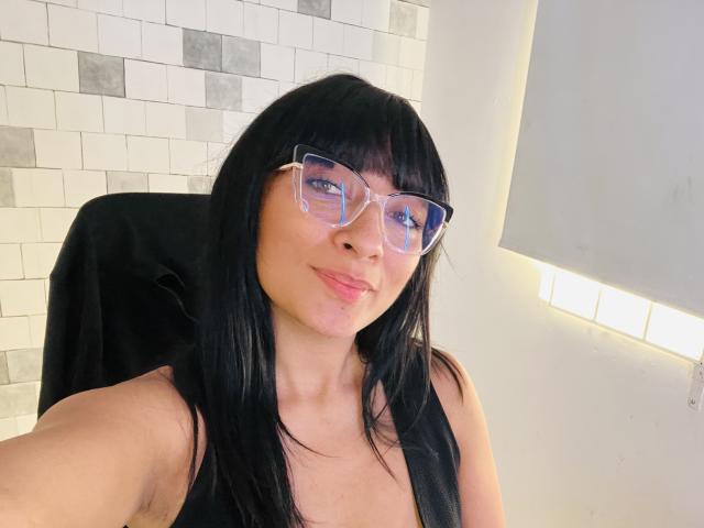 AmelineRousseau has long black hair and wears clear glasses. She is smiling slightly with pink lips. She is in a black chair and has a fair skin tone. The background has white and gray tiles. The lighting is bright from the right. Her face is centered, and she looks directly at the camera. The image is a selfie.