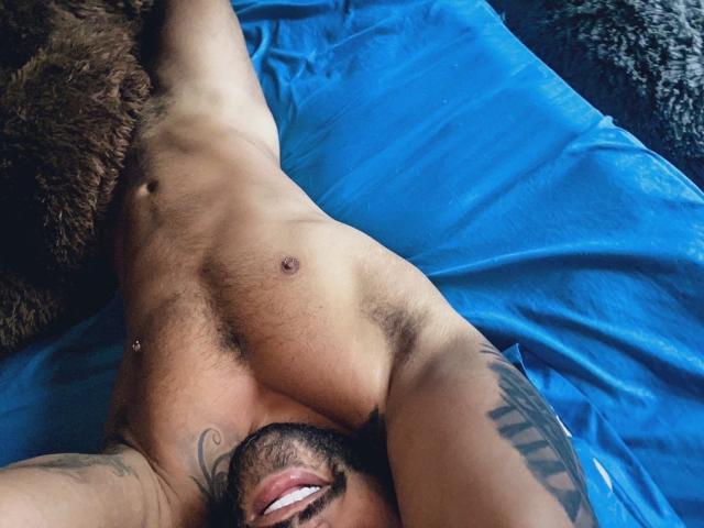 DammianSafado, sem camisa, musculoso, tatuado, com os braços atrás da cabeça, olhos castanhos, barba, brincos, pênis flácido em uma sunga bege, peito peludo, axilas, olhando para a câmera, luz do sol, cama..