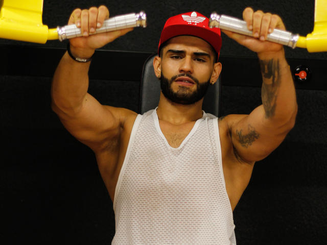 Dammiansafado1, muscular and tan, with a trimmed beard, wears a red Adidas cap and a white mesh tank top. He's holding yellow dumbbells overhead, arms fully extended. Tattoos on his right bicep and left forearm. Black textured wall behind him. His intense, focused expression. Bright lighting. Slightly shadowed face and upper body. Strong, defined muscles and confident posture. Photo focuses on upper body and face.