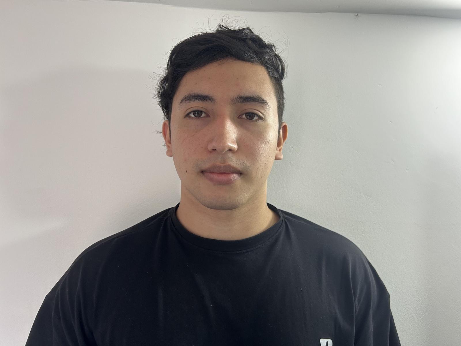Photograph of PruebaMan. PruebaMan has medium-length, slightly wavy black hair, brown eyes, and a small mole on his right cheek. He's wearing a simple black t-shirt. His skin is light brown, and he has a neutral expression. The background is a plain white wall, and the lighting is even. PruebaMan's face is centered, and his t-shirt contrasts with the white wall. The image is clear, focusing on PruebaMan's upper body.